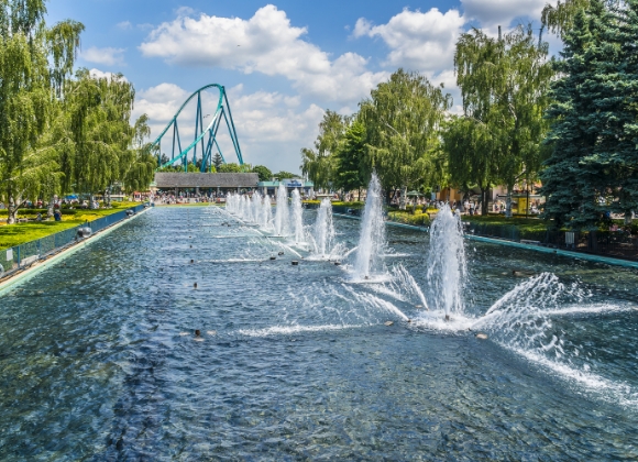 A large pond with several fountains spraying water and trees lining the sides. In the distance is a teal roller coaster.