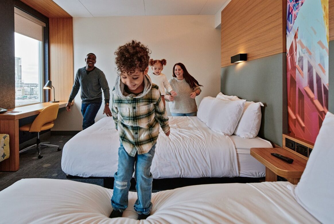 Little boy jumping on bed in a hotel room with his parents and younger sibling watching behind him.