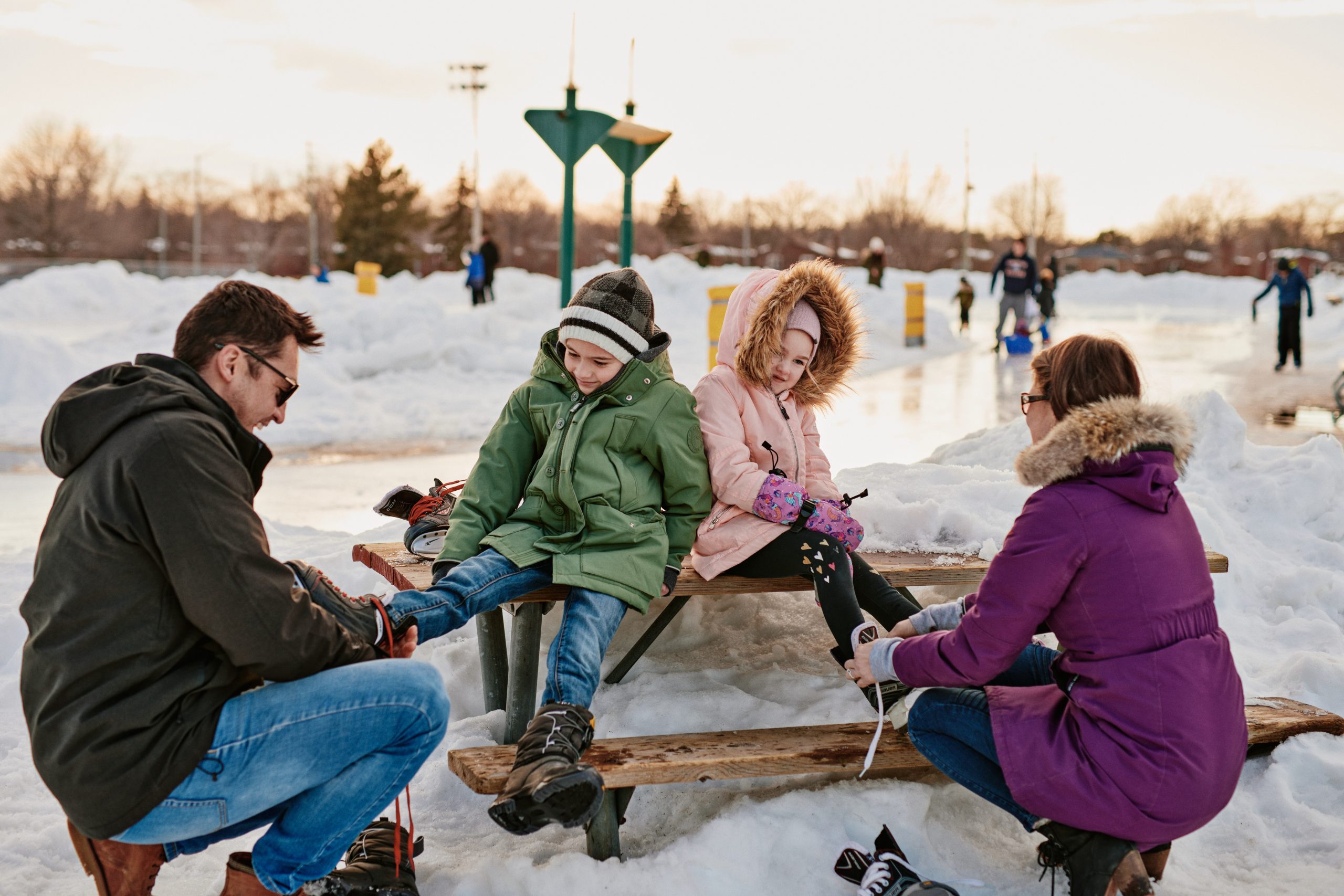 A family of four sitting on a snowy bench, with the parents putting skates on their children.