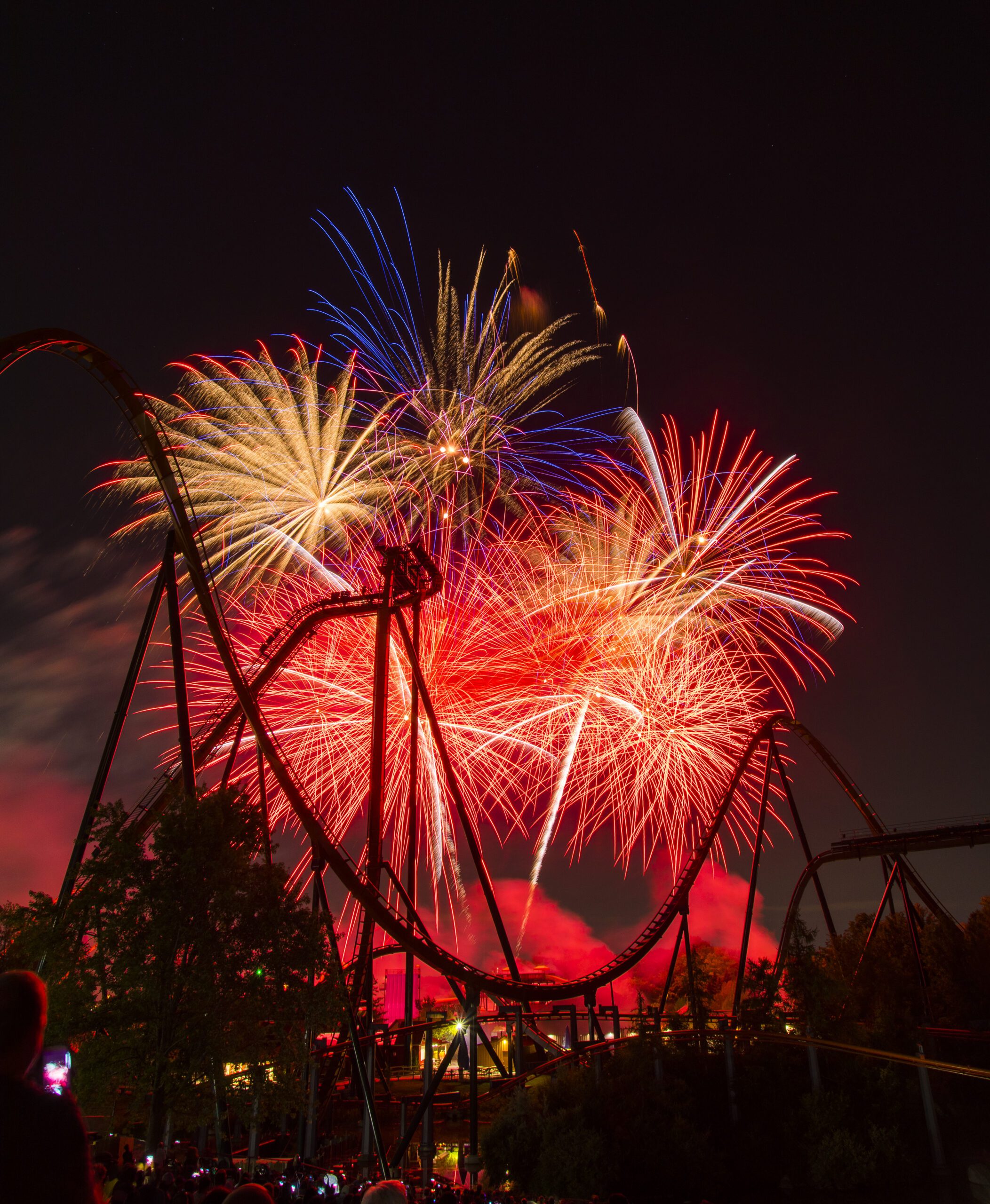 Red and yellow fireworks in a dark sky with the silhouette of a roller coaster in the forefront.
