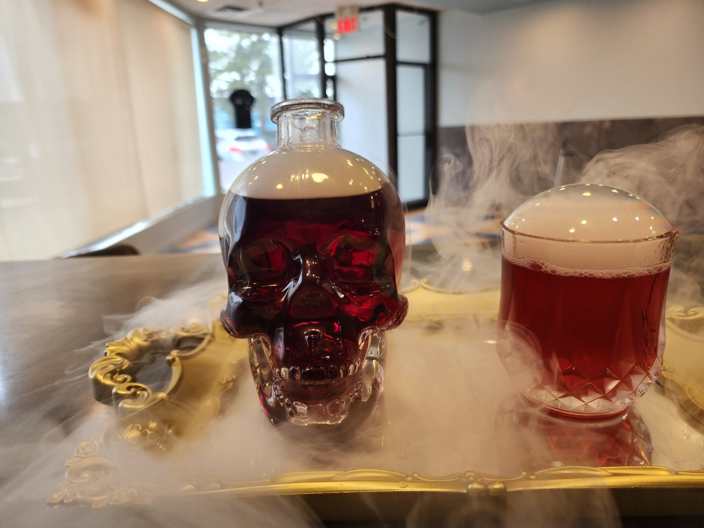 A clear glass skull container filled with a dark liquid, sitting beside a beer with smoke all around.