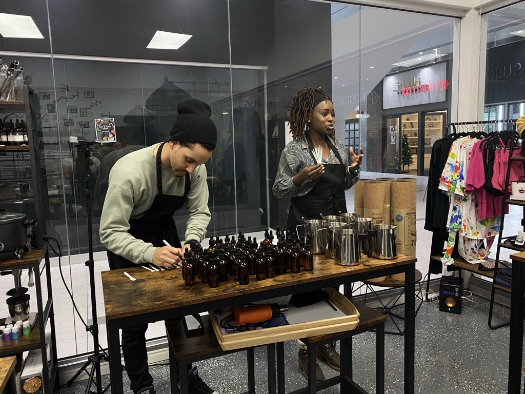 A woman speaking with a man by her side assisting and a table of candle making products before them.