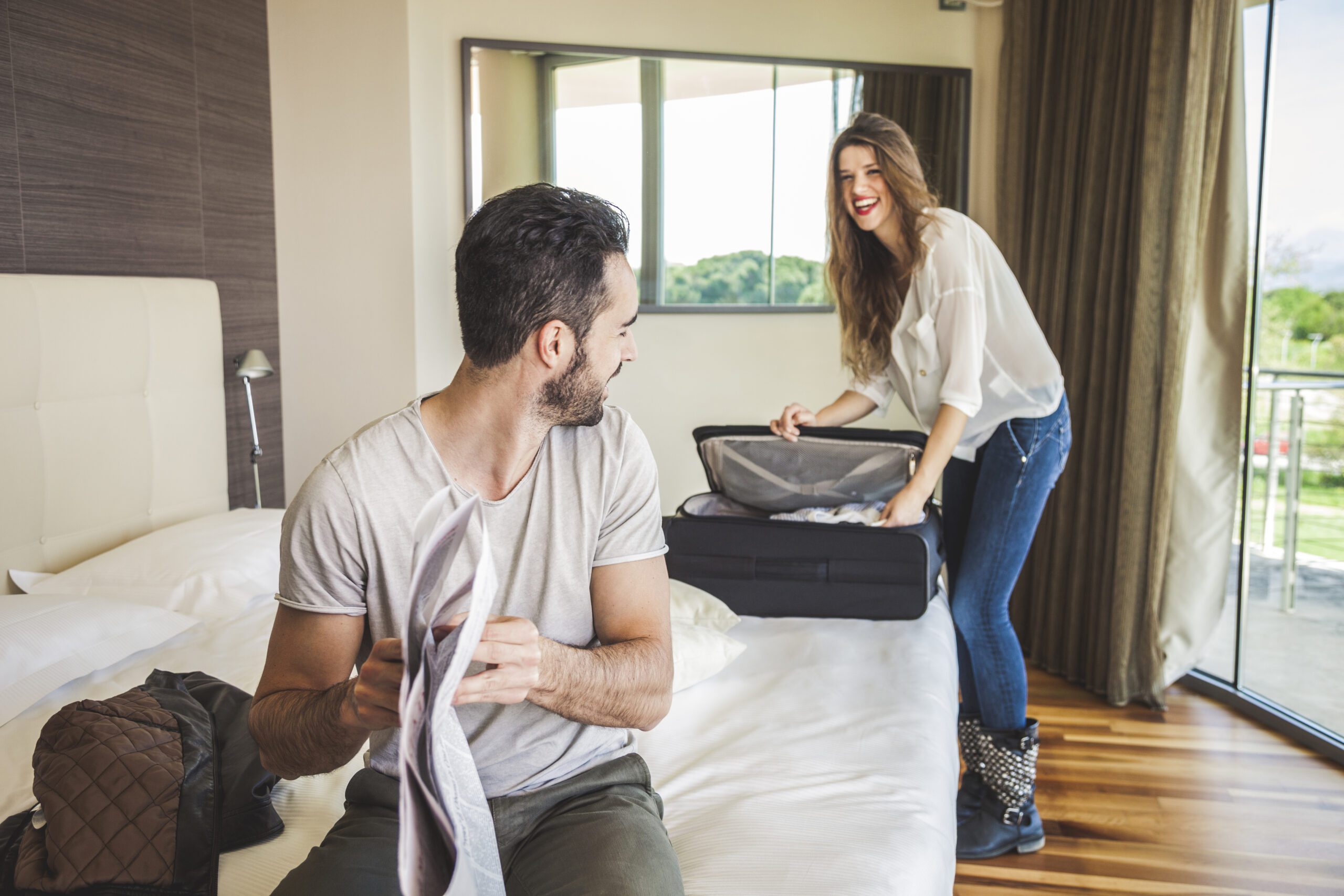 A man sitting on a bed looks back at his female partner who is unpacking a suitcase and laughing.