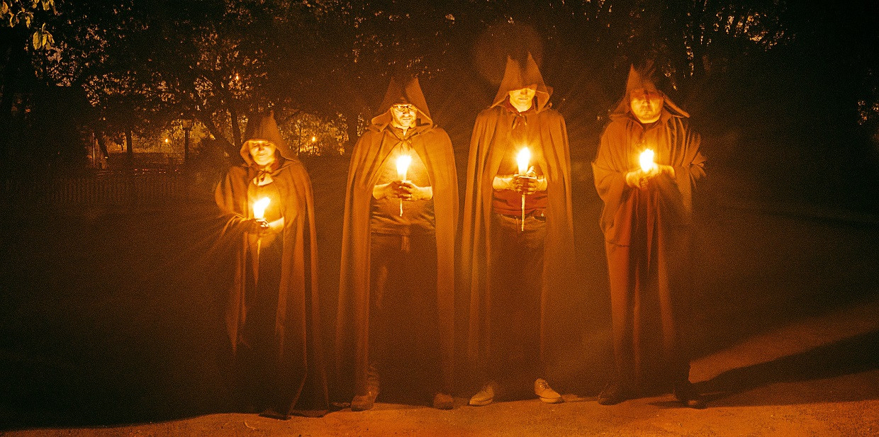 Four hooded figures in cloaks holding lanterns in the darkness