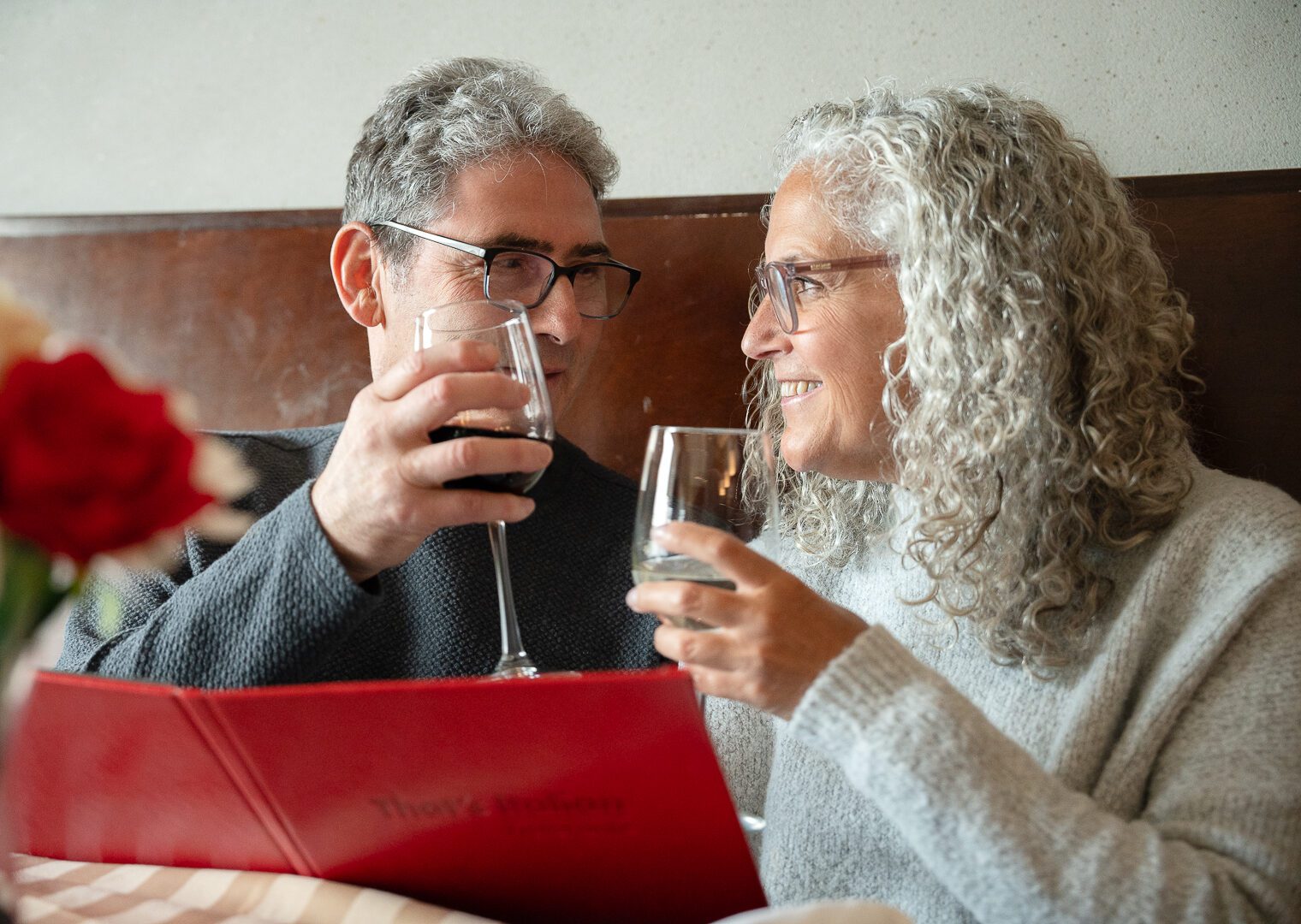 Image of two adults sitting close to each other at a restaurant with glasses of wine in their hand as they look at each other lovingly