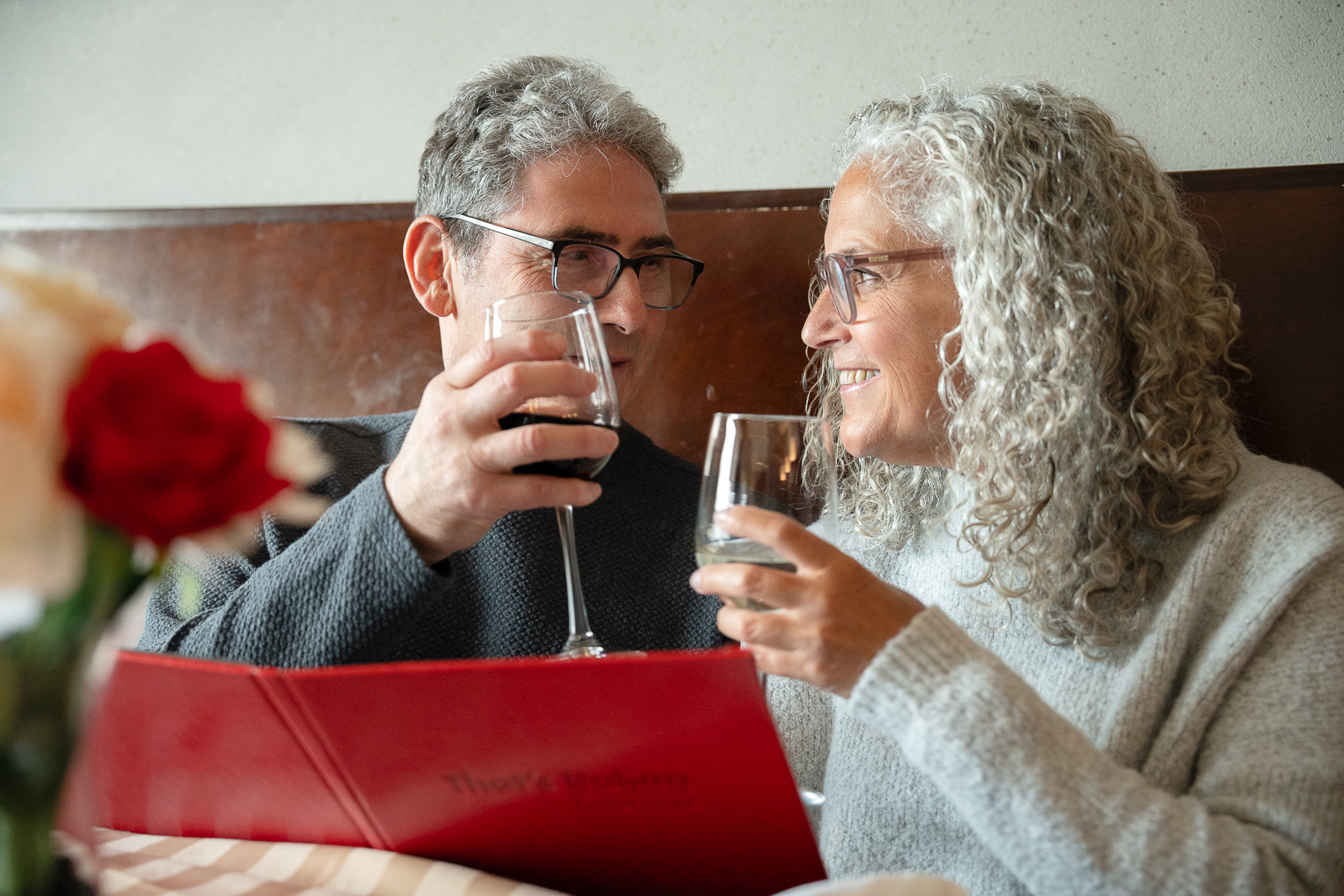 Image of two adults sitting close to each other at a restaurant with glasses of wine in their hand as they look at each other lovingly