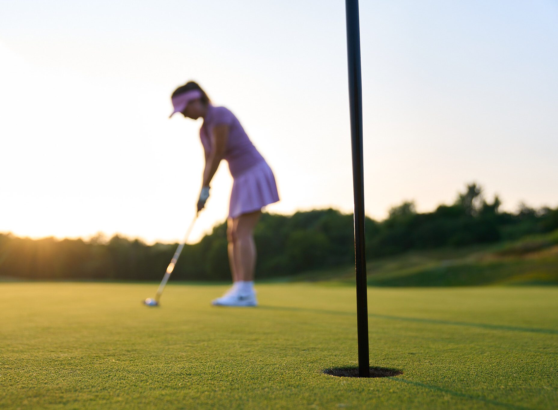 A woman on a golf green putting a golf ball into a hole, the sun is setting in the background
