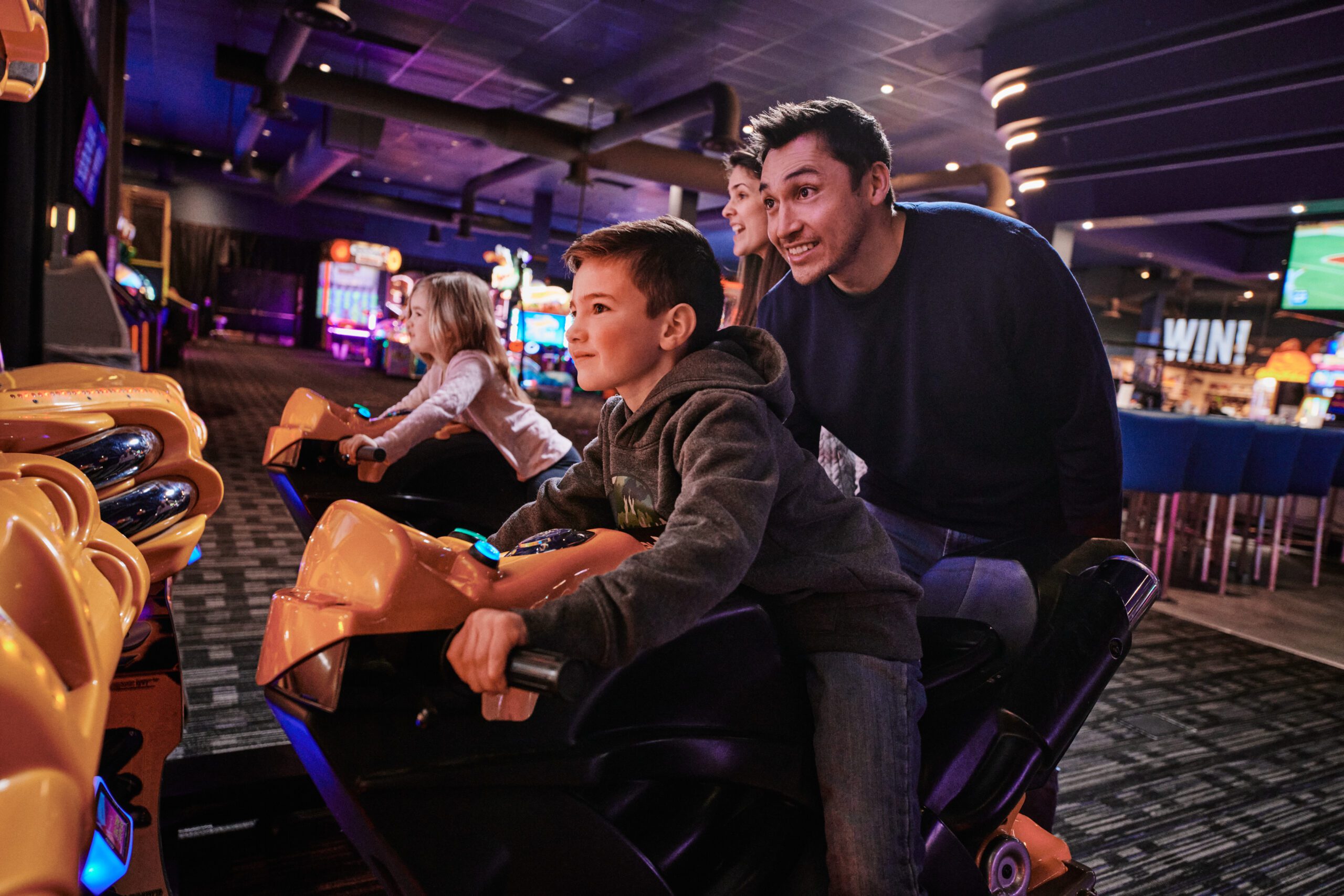 Two children racing on arcade motorcycle bikes, the father is standing behind watching