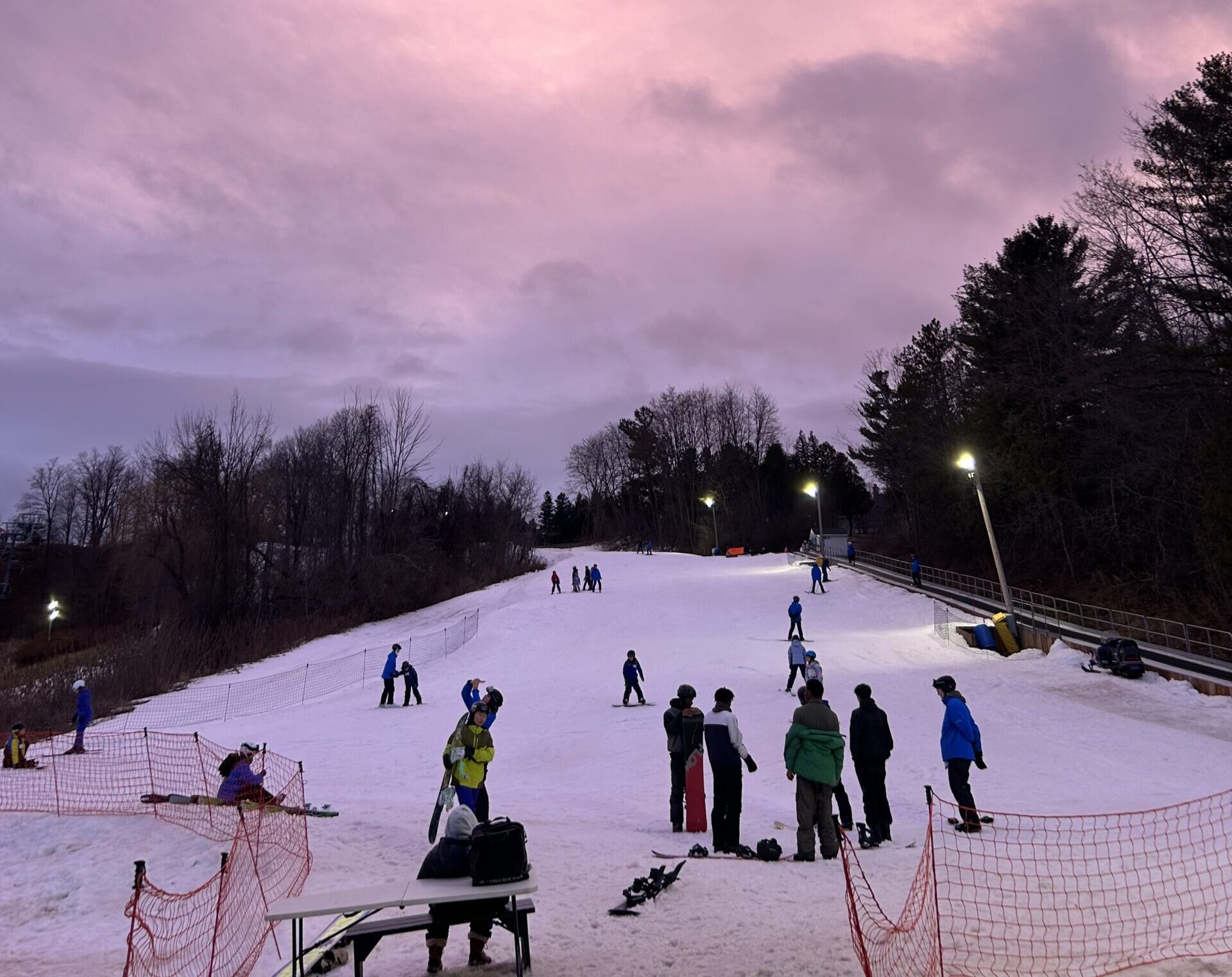 Image taken at the bottom of a ski hill looking up towards the top with people skiing down and waiting in line for the chair lift. The sky is pink and purple from the setting sun.