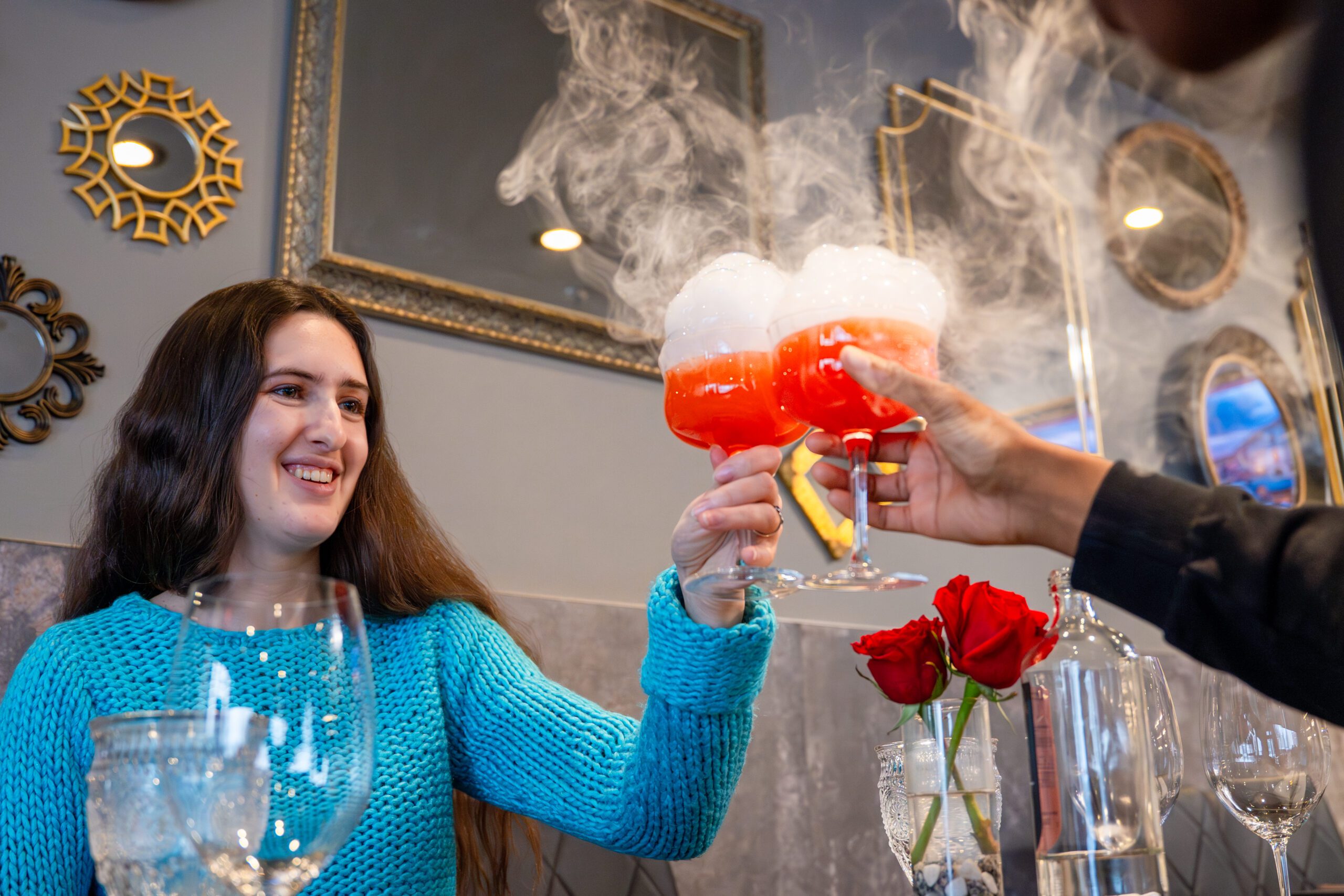 Image of a person clinking glasses with another person off camera. The drinks are red, topped with foam, and have smoke coming out from them.