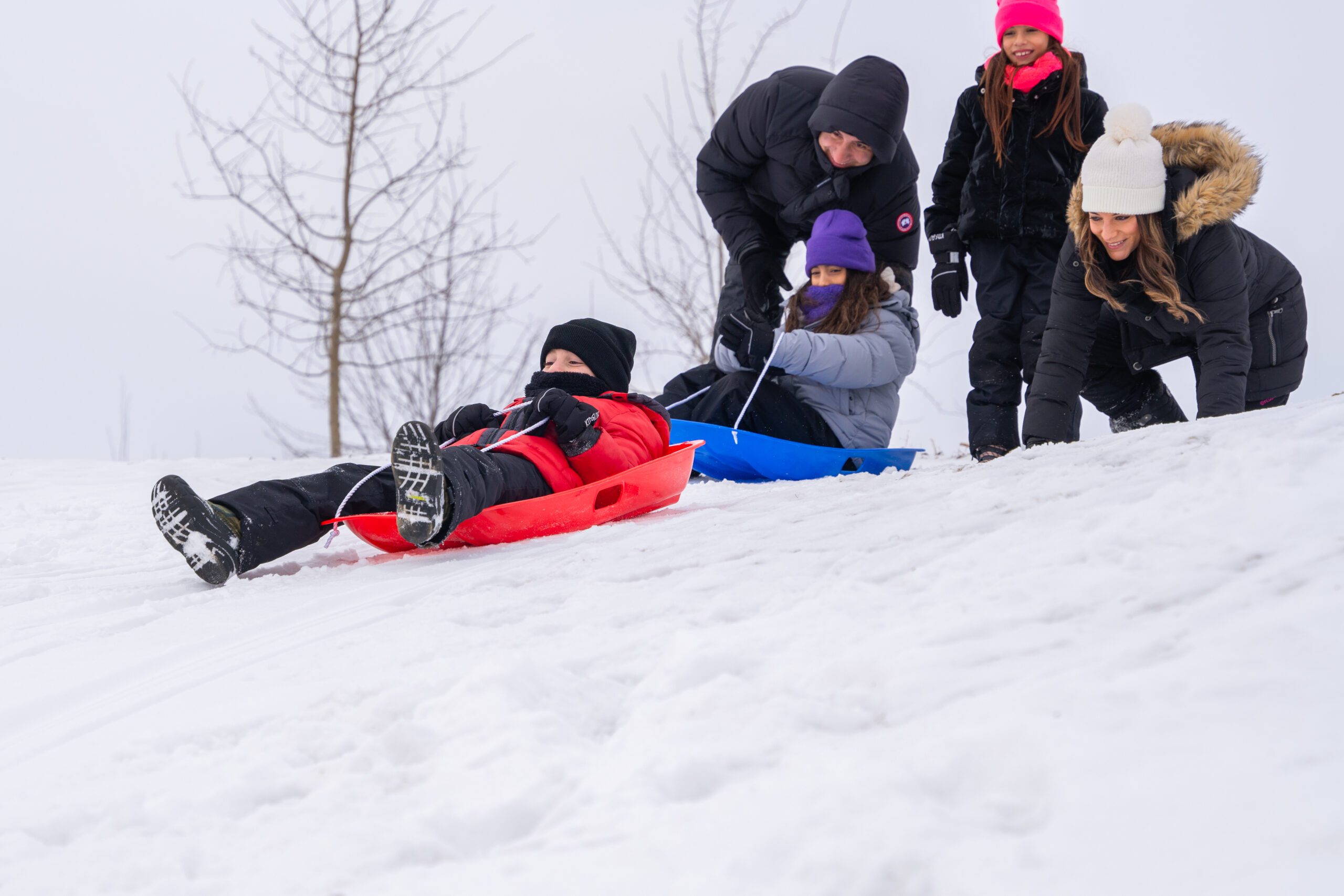 Image of two children sitting on sleds at the top of a snowy hill with two adults and a third child pushing them