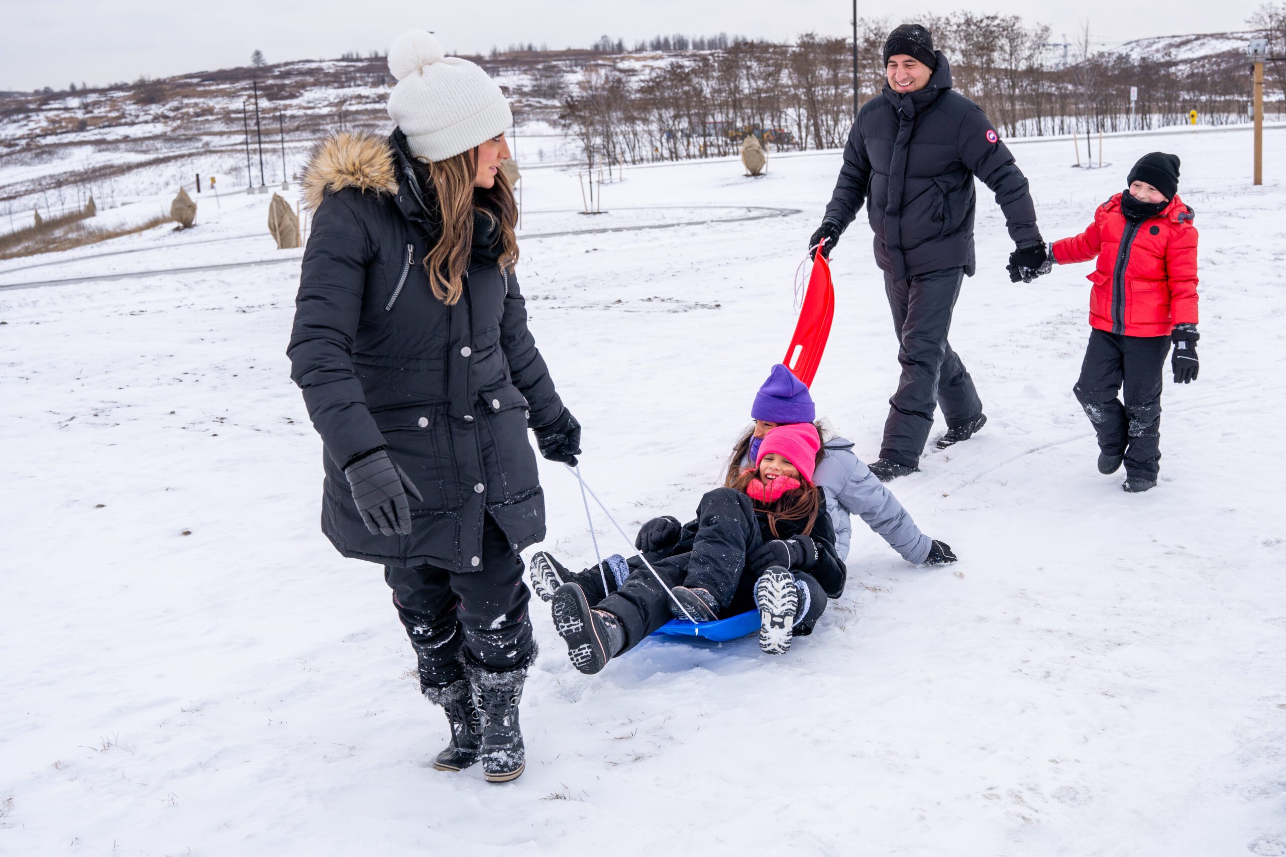 A woman pulling her two kids on a blue plastic sled, the father is following behind