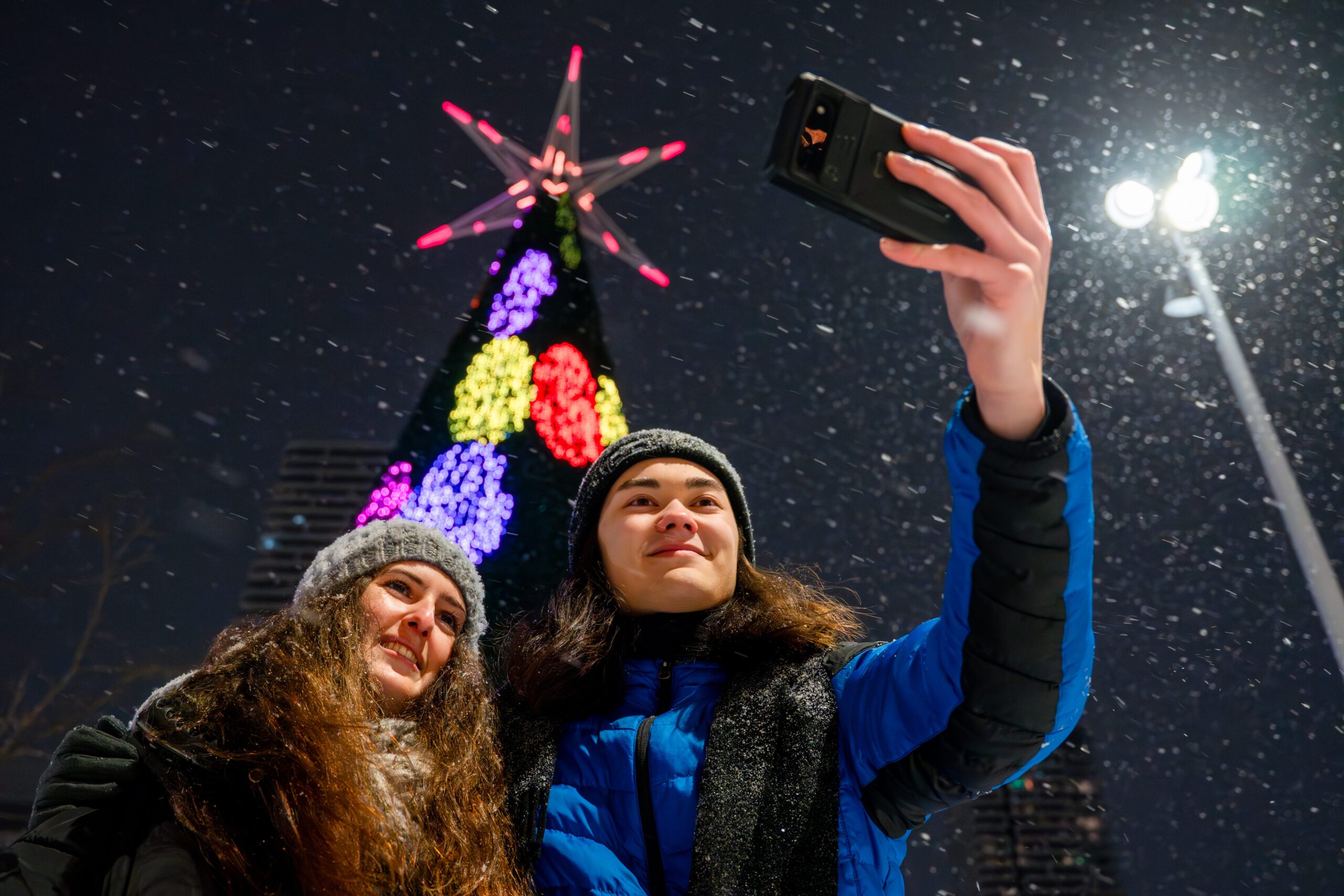 Image of two people taking a selfie in front of a large, illuminated Christmas tree outdoors at nighttime