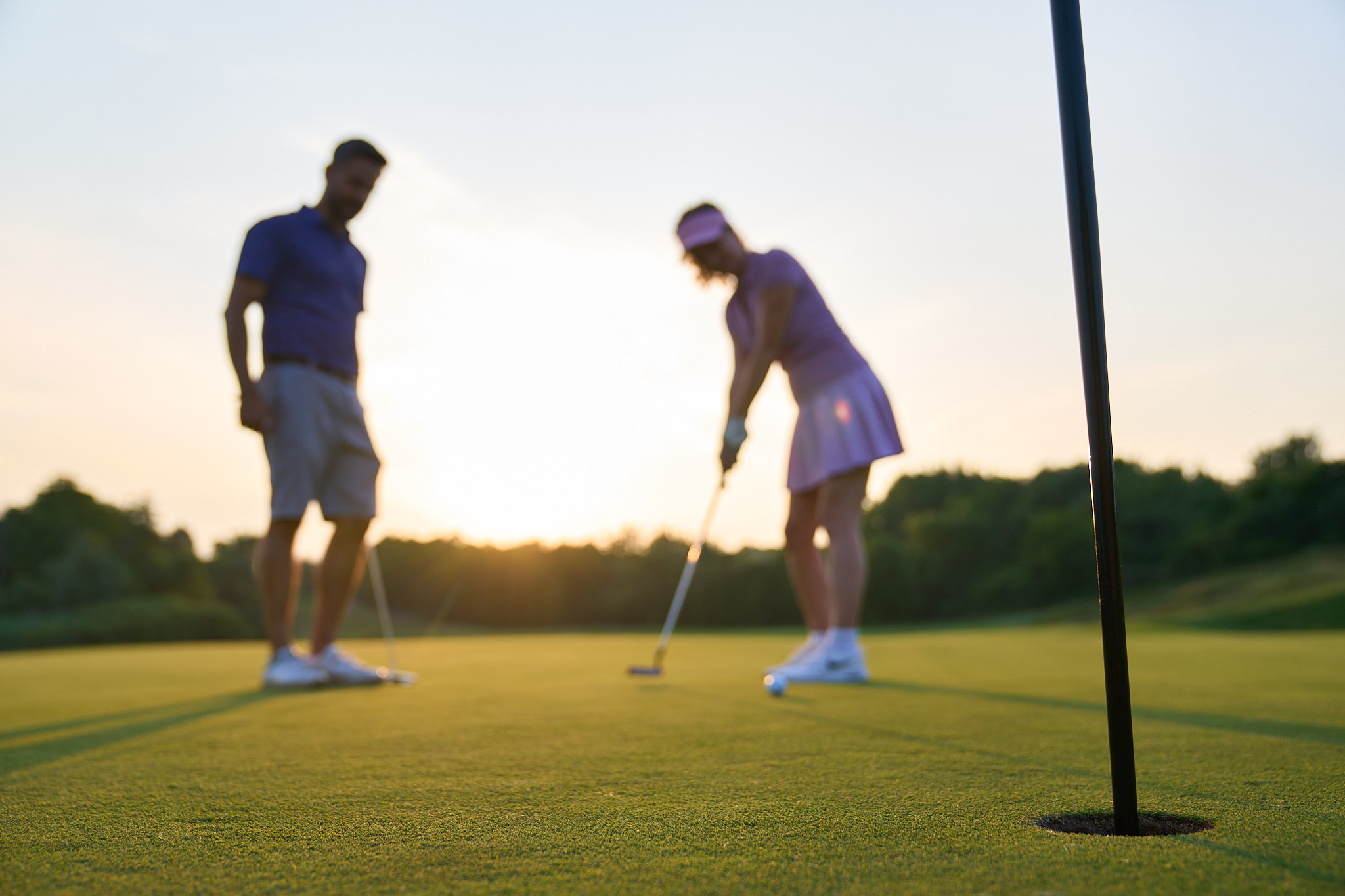 Image of two people standing on a golf course in front of a sunset. One of them is putting.