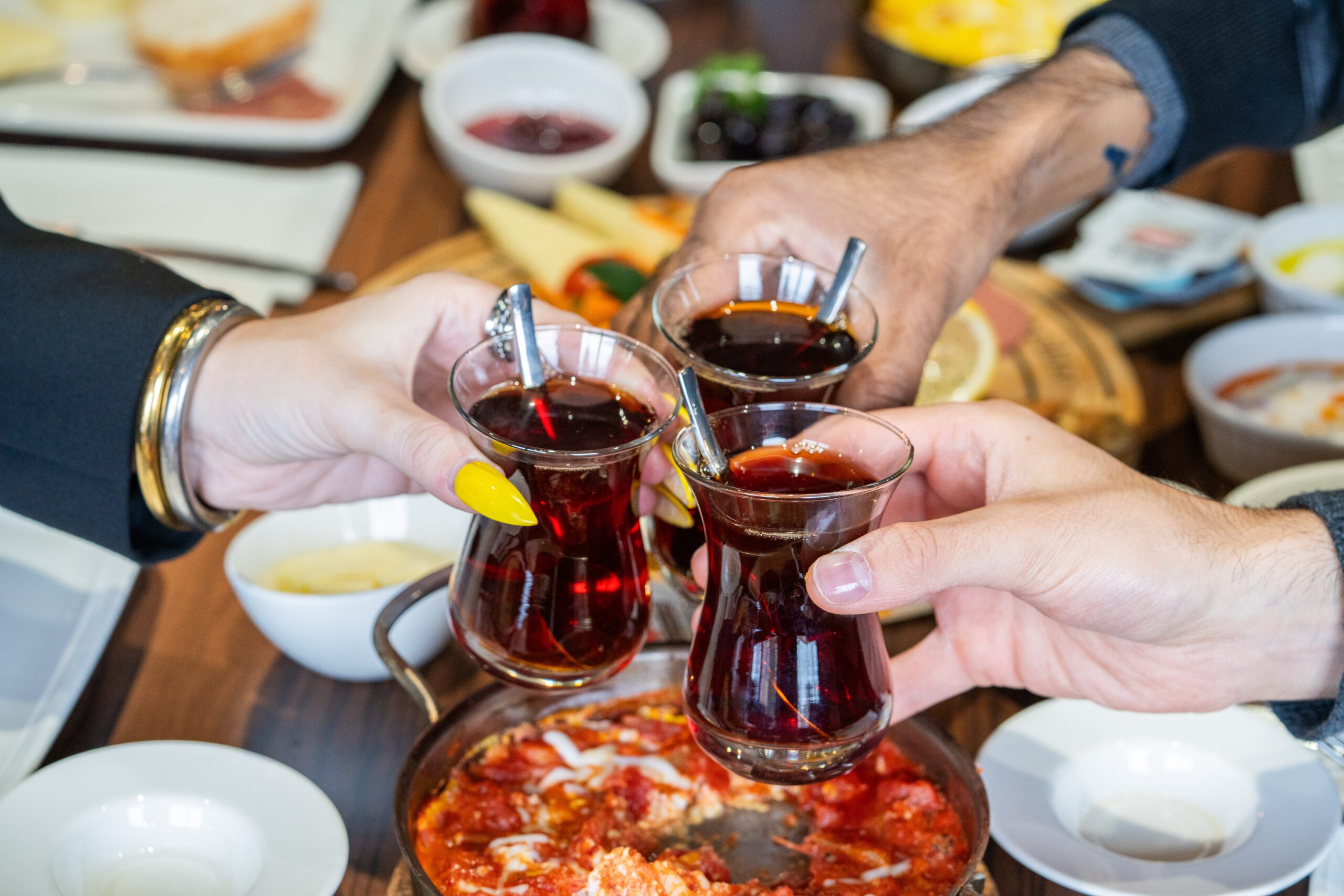 Image of three hands holding cups of Turkish tea, clinking glasses over a table full of brunch food and sweets