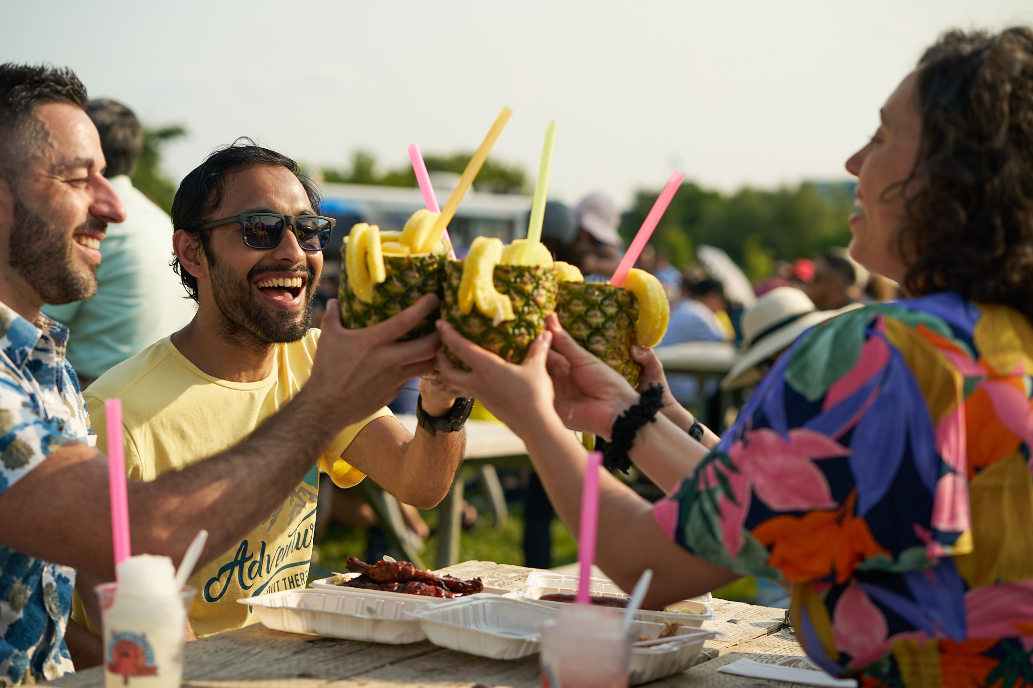 Image of a group of friends sitting at a table at a festival that are doing a toast using drinks served in a pineapple